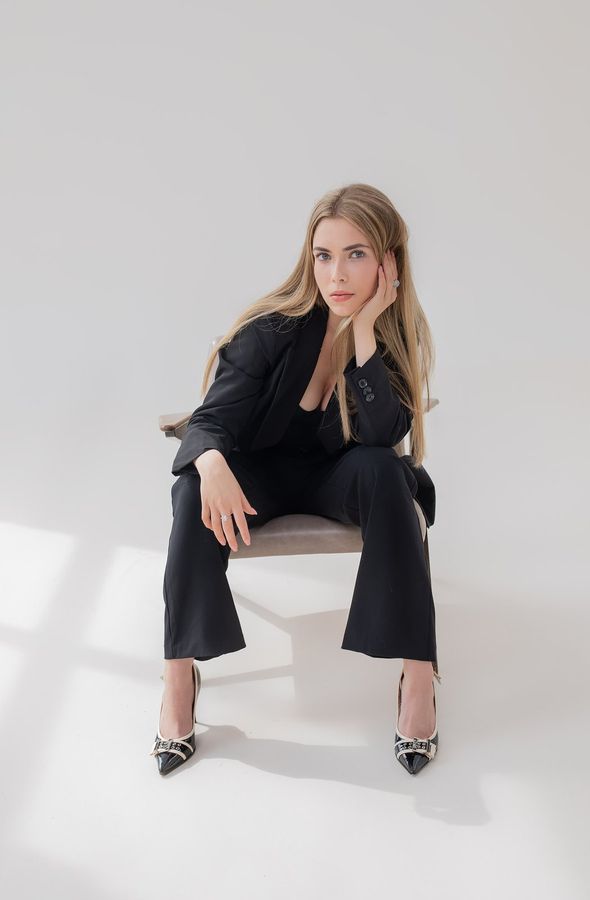 Білява дівчина в чорному брючному костюмі сидить на стільці, лікоть на коліні, щока на долоні